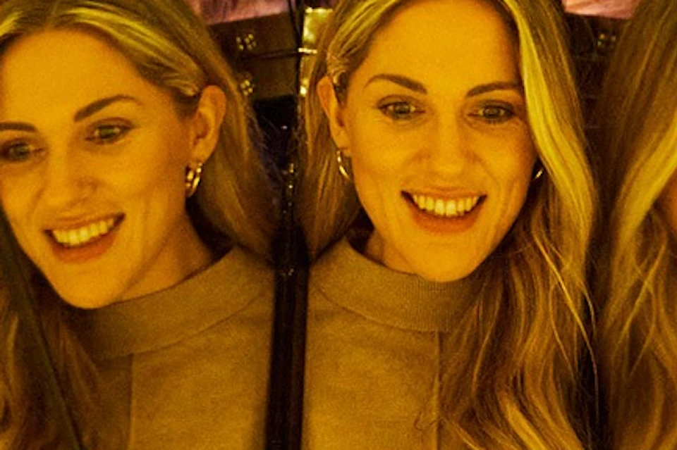A woman with long blond hair is reflected multiple times in a mirror installation with warm lighting and green lights visible.