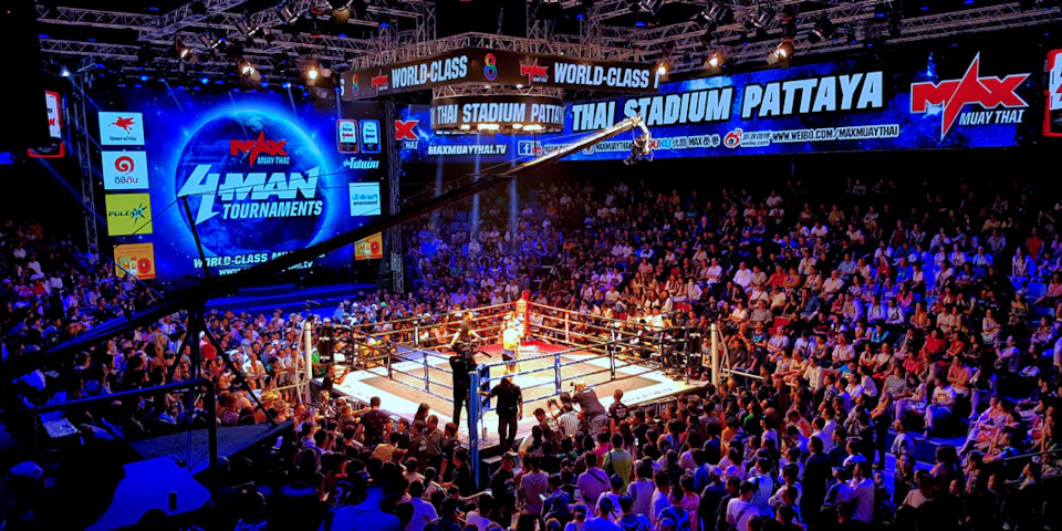 A crowded indoor stadium with a boxing ring in the center, surrounded by spectators and illuminated by bright lights.