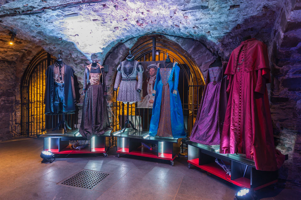 Mannequins display ornate historical costumes in a dimly lit stone room, with an arched doorway and barred gate in the background.