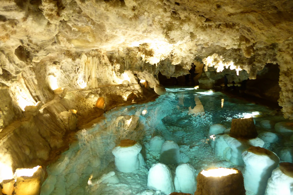 A cave with illuminated stalactites and stalagmites, and a clear blue underground pool with reflections on the water surface.