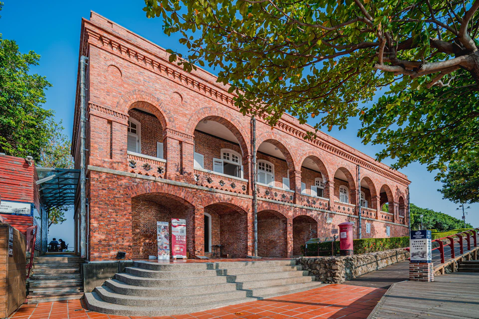 A historic red-brick building with arched windows, a staircase leading to the entrance, and leafy trees in the foreground.