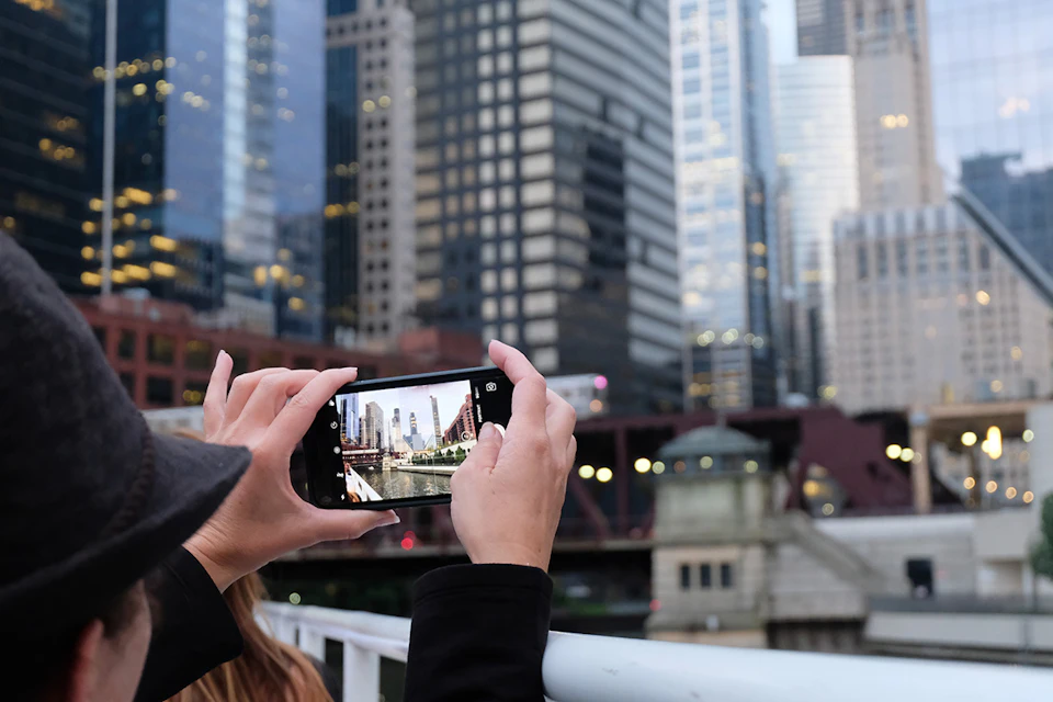A person takes a photo of a city skyline with a smartphone, capturing tall buildings and a bridge in the background.