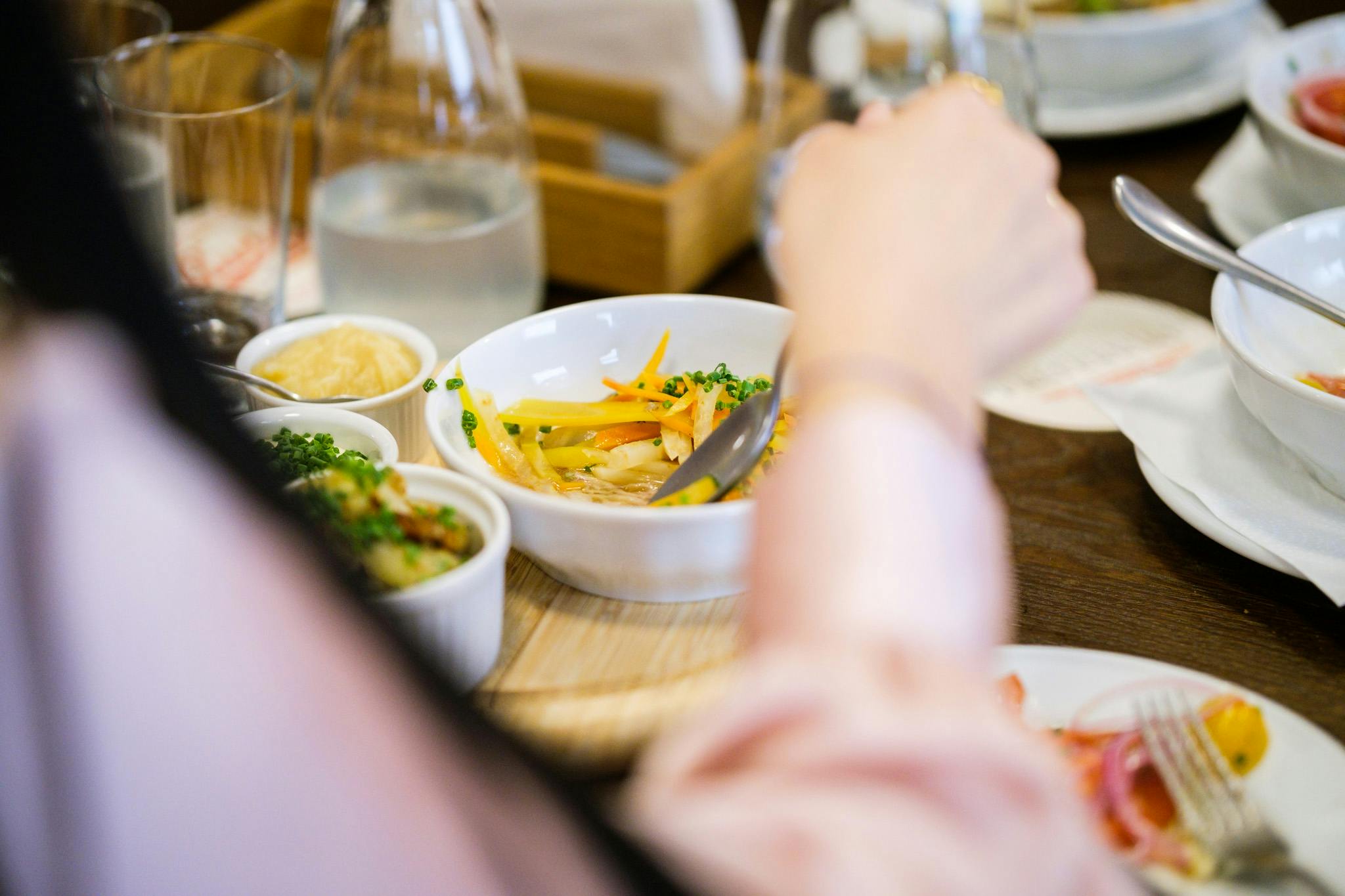 Osoba držící lžíci nad miskou polévky se zeleninou. Na stole je několik malých pokrmů a láhev na vodu.