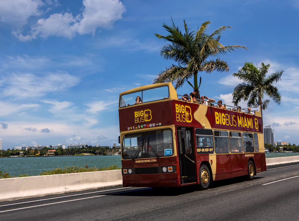 A double-decker tour bus with "Big Bus Miami" branding drives on a road near water and palms, with passengers on the top deck.