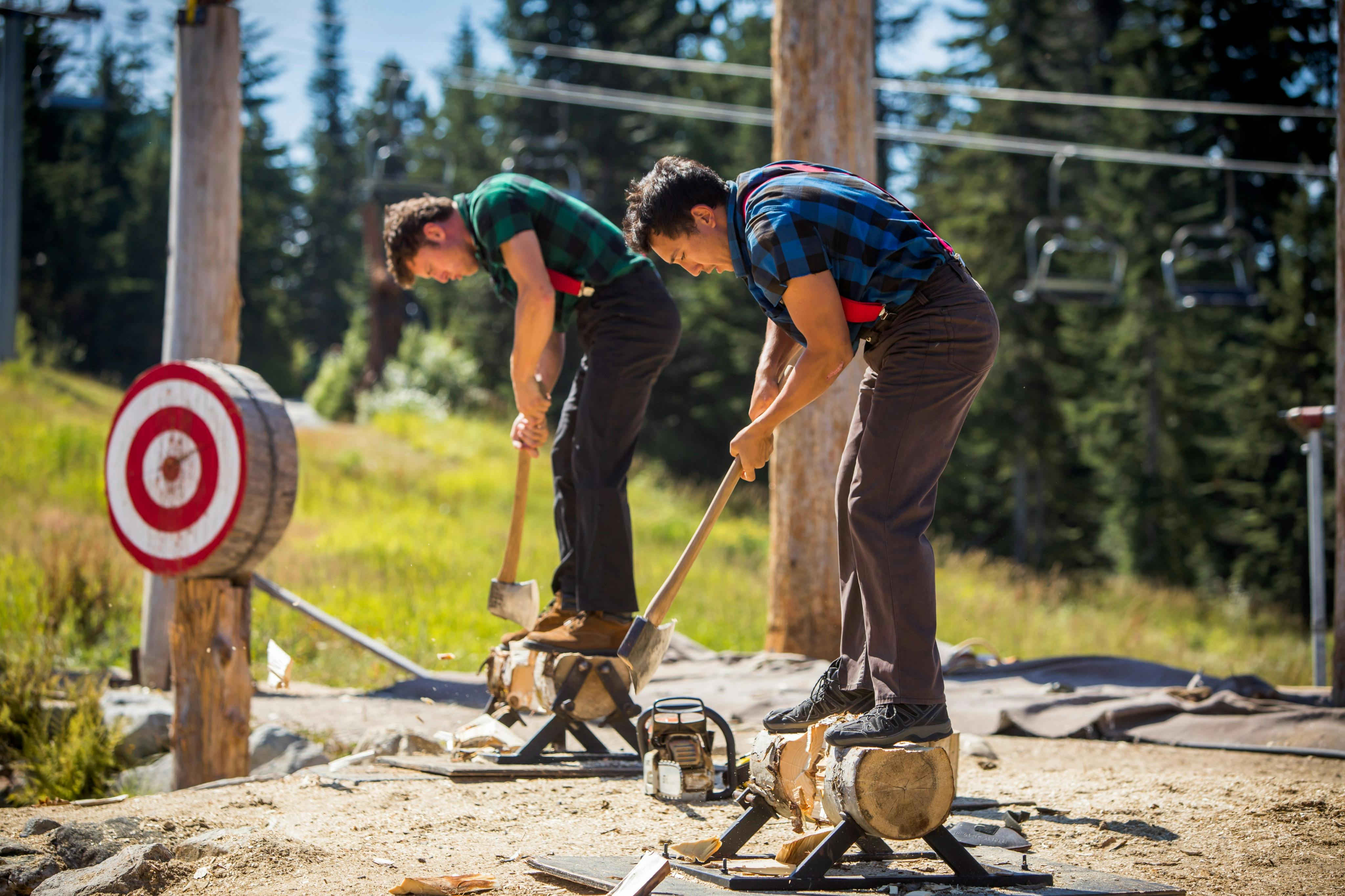 격자무늬 셔츠와 멜빵을 입은 두 사람이 나무와 탁 트인 하늘로 둘러싸인 야외에서 나무를 자르고 있습니다.