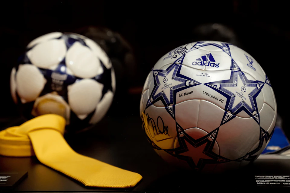 A signed Adidas soccer ball with blue star patterns, featuring the teams AC Milan and Liverpool FC, displayed on a dark surface.
