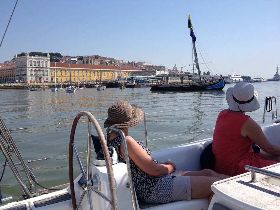 Two people wearing hats sit on a boat, looking at a sailboat and a yellow building across the water under a clear sky.