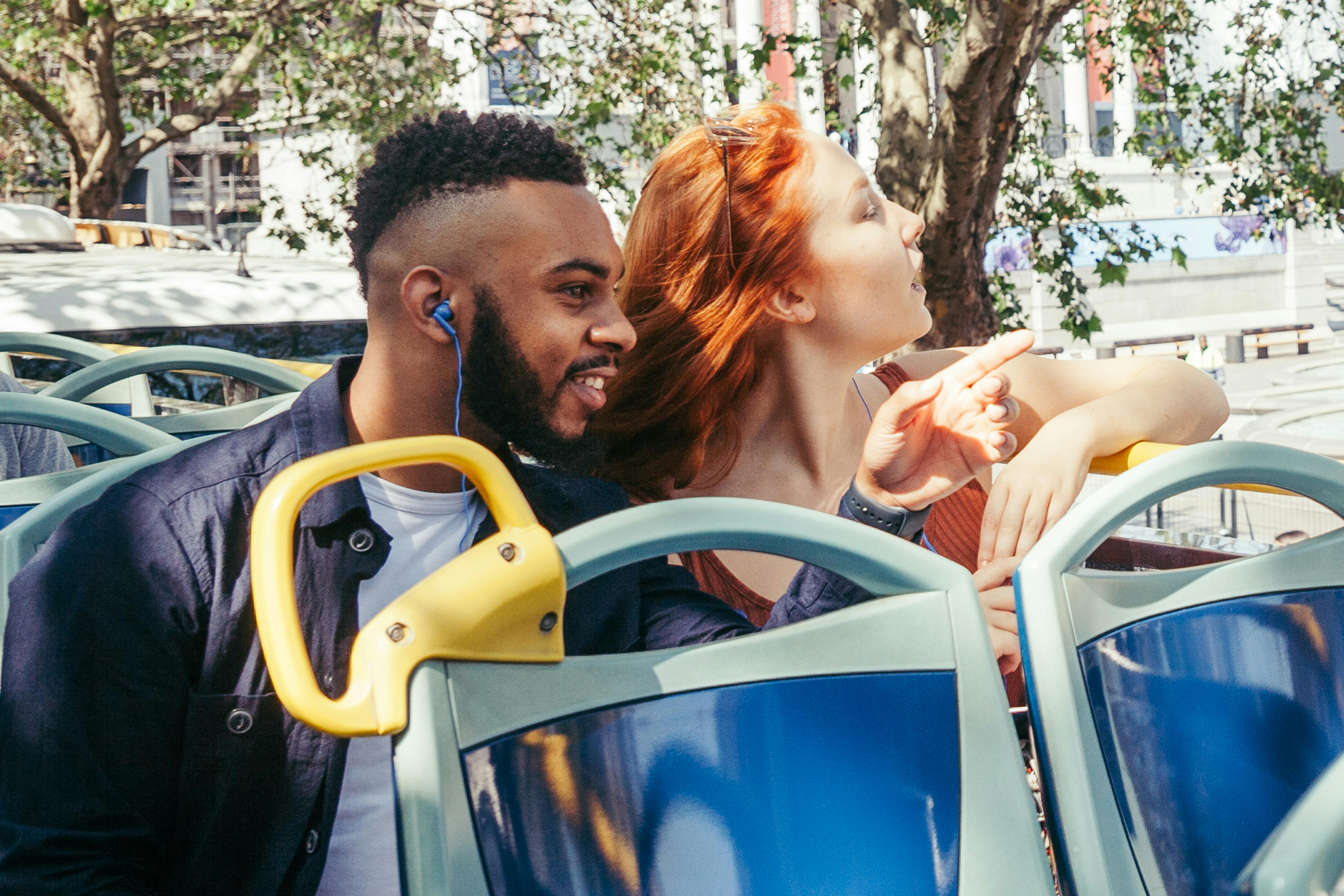 Two people ride in an open-top bus, enjoying the view and pointing at something. Trees and buildings are in the background.