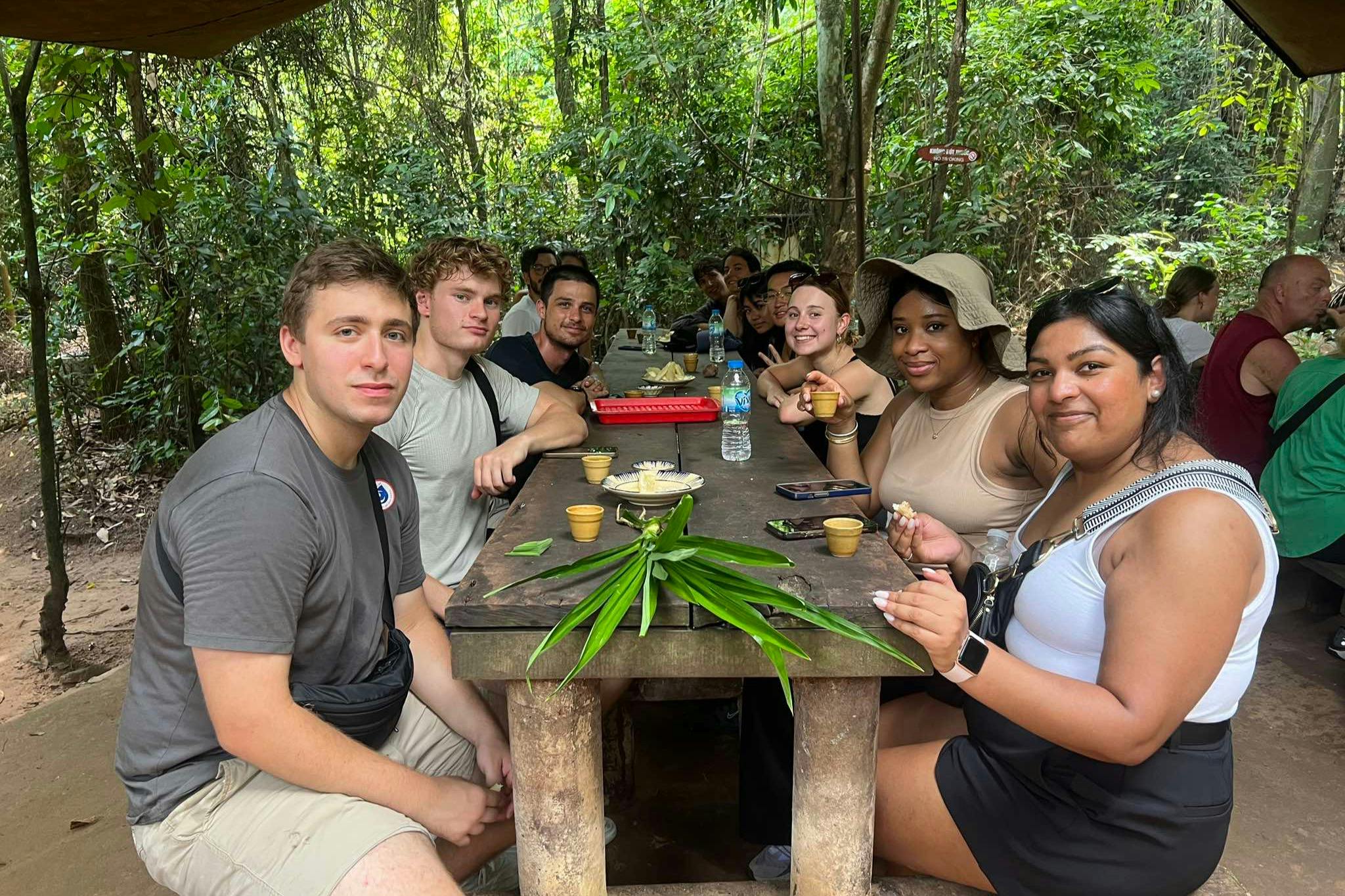 A group of people sitting at a wooden table outdoors, surrounded by dense forest, with food and drinks on the table.