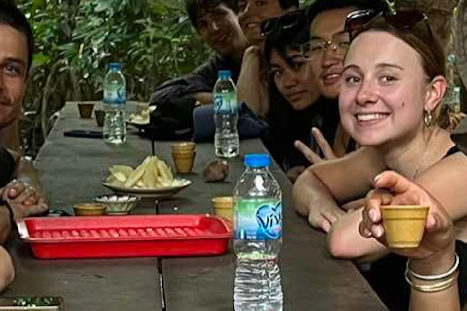 A group of people sitting at a wooden table outdoors, surrounded by dense forest, with food and drinks on the table.