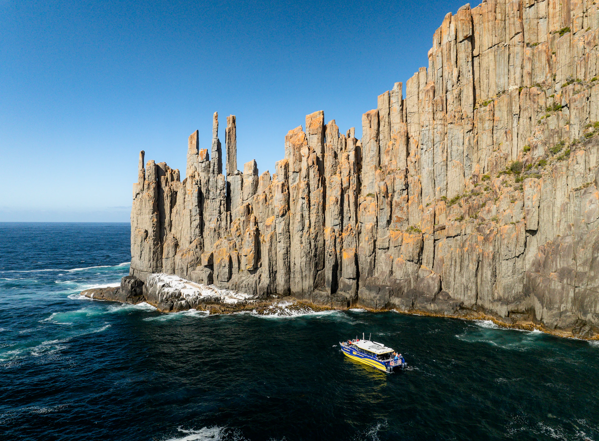 A small boat sails near a tall, rugged cliff face by the ocean under a clear blue sky.