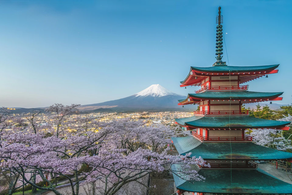A pagoda with cherry blossoms in the foreground and Mount Fuji in the background under a clear blue sky.