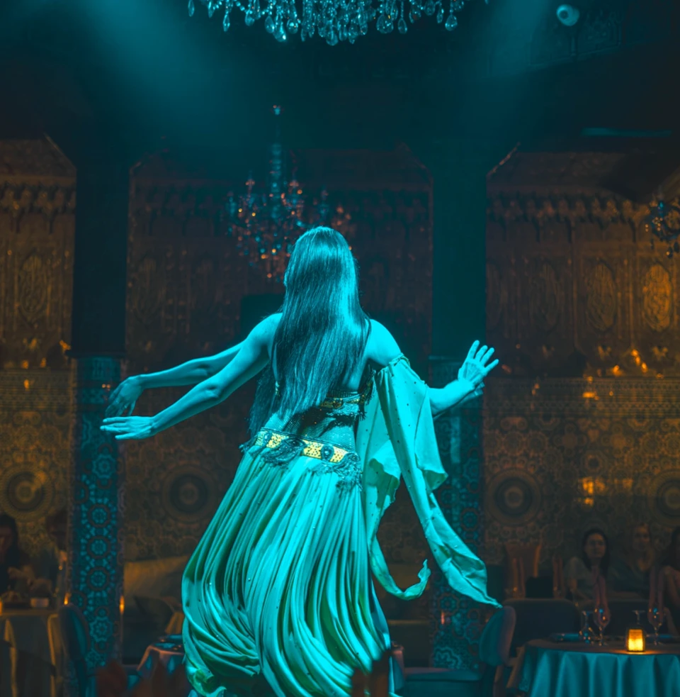 A woman in a flowing green dress dances gracefully in a dimly lit, ornately decorated room with chandeliers.