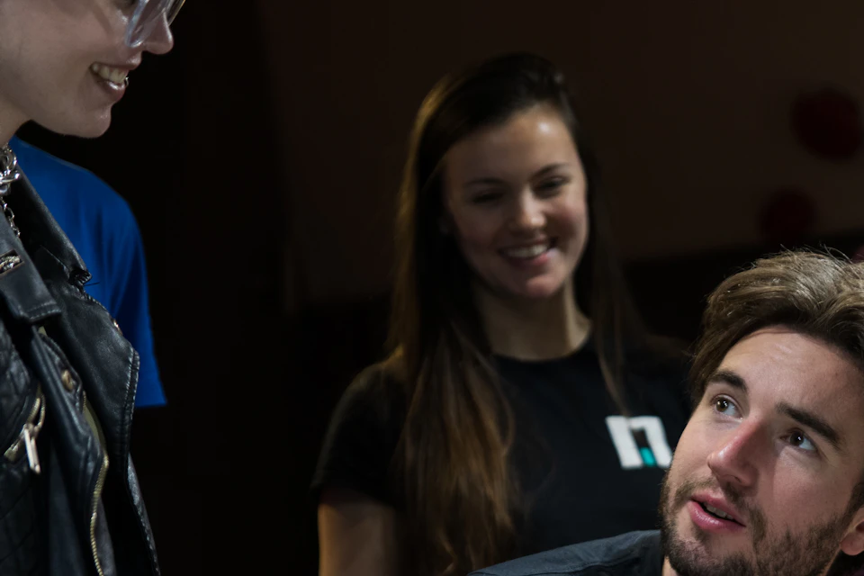 Three people interacting indoors, with one person seated, looking up, and two standing. Casual attire, dark background.