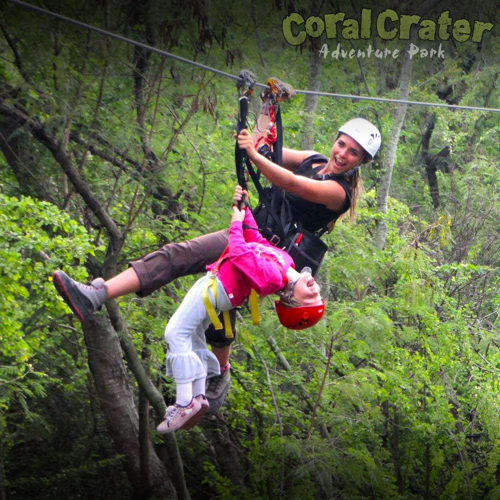 Δύο άνθρωποι με φερμουάρ σε ένα δάσος, ένας ενήλικας υποστηρίζει ένα παιδί που κρέμεται ανάποδα. Κείμενο: "Coral Crater Adventure Park."