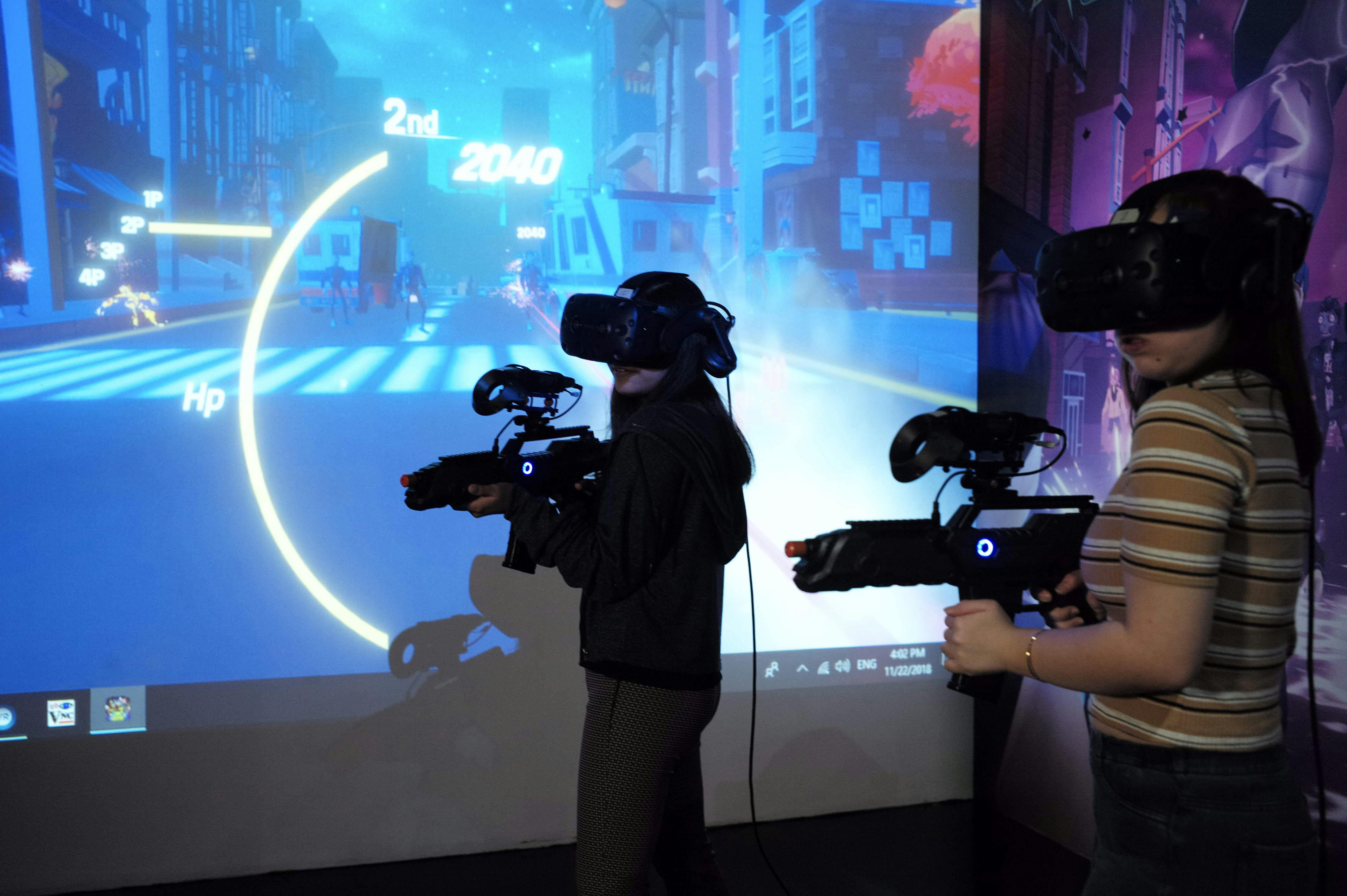 Deux personnes portant des casques VR et tenant des pistolets de jeu devant un grand écran de jeu vidéo futuriste.