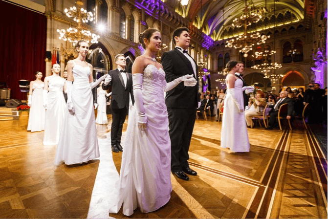 Άτομα με επίσημη ενδυμασία που συμμετέχουν σε μια εκδήλωση σε αίθουσα χορού, με πολυελαίους και τοξωτά ταβάνια στο παρασκήνιο.