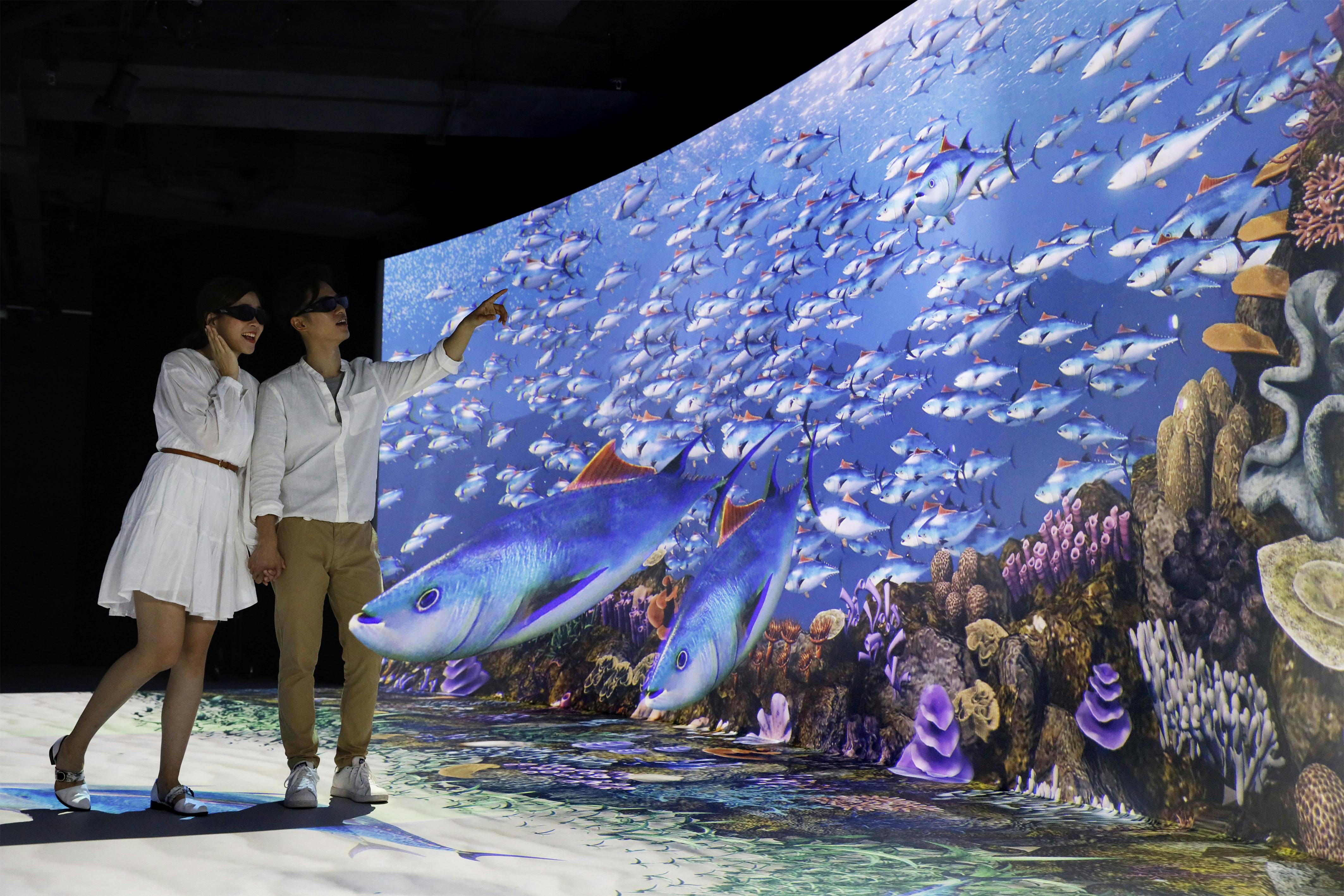 Двое людей в 3D-очках любуются большой, захватывающей демонстрацией подводной сцены с рыбой и кораллами.