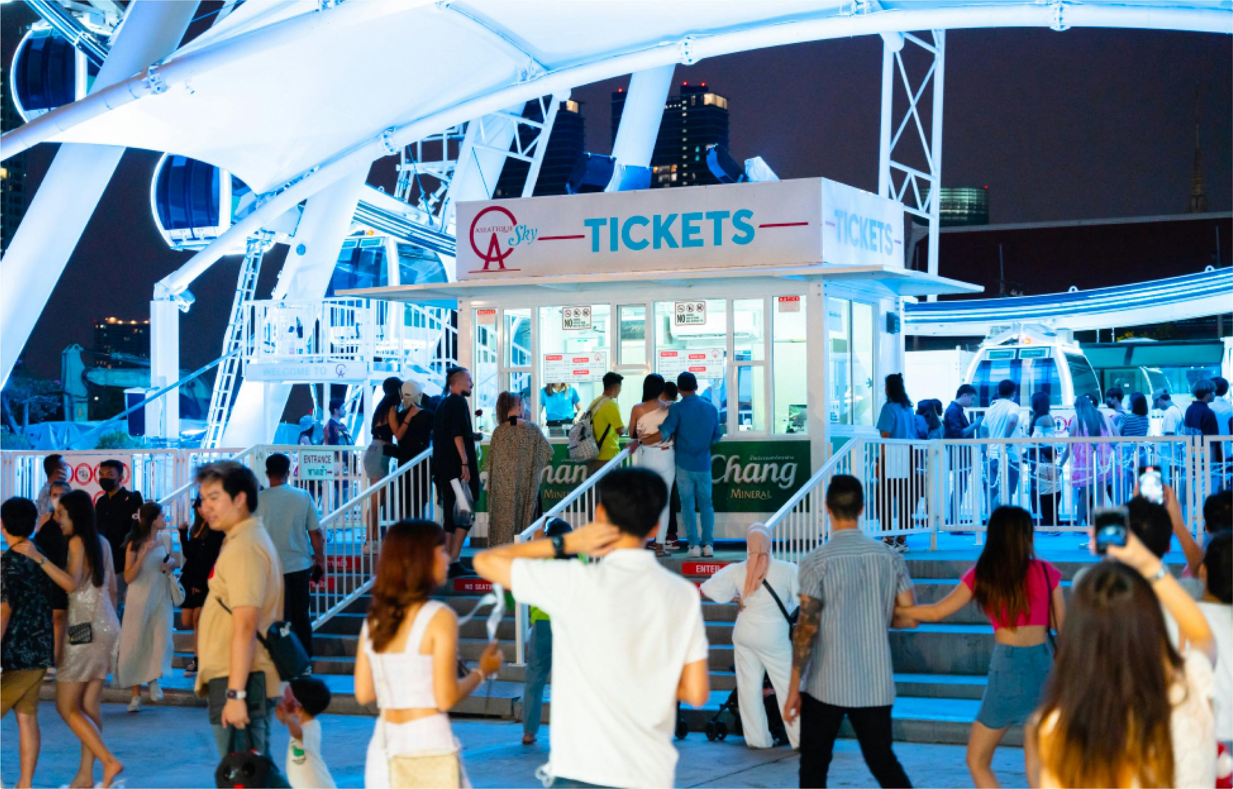 People gathering near a ticket booth illuminated with blue lights in an outdoor setting with a ferris wheel structure above.