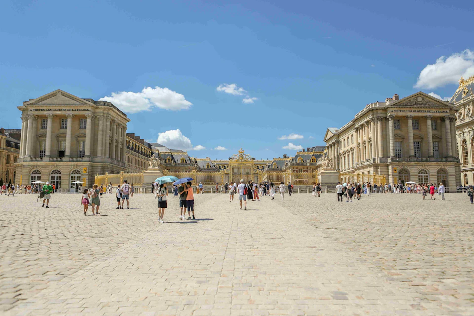 Tourists walk and gather in a large cobblestone courtyard in front of the ornate gates of a grand historical building under a clear blue sky.