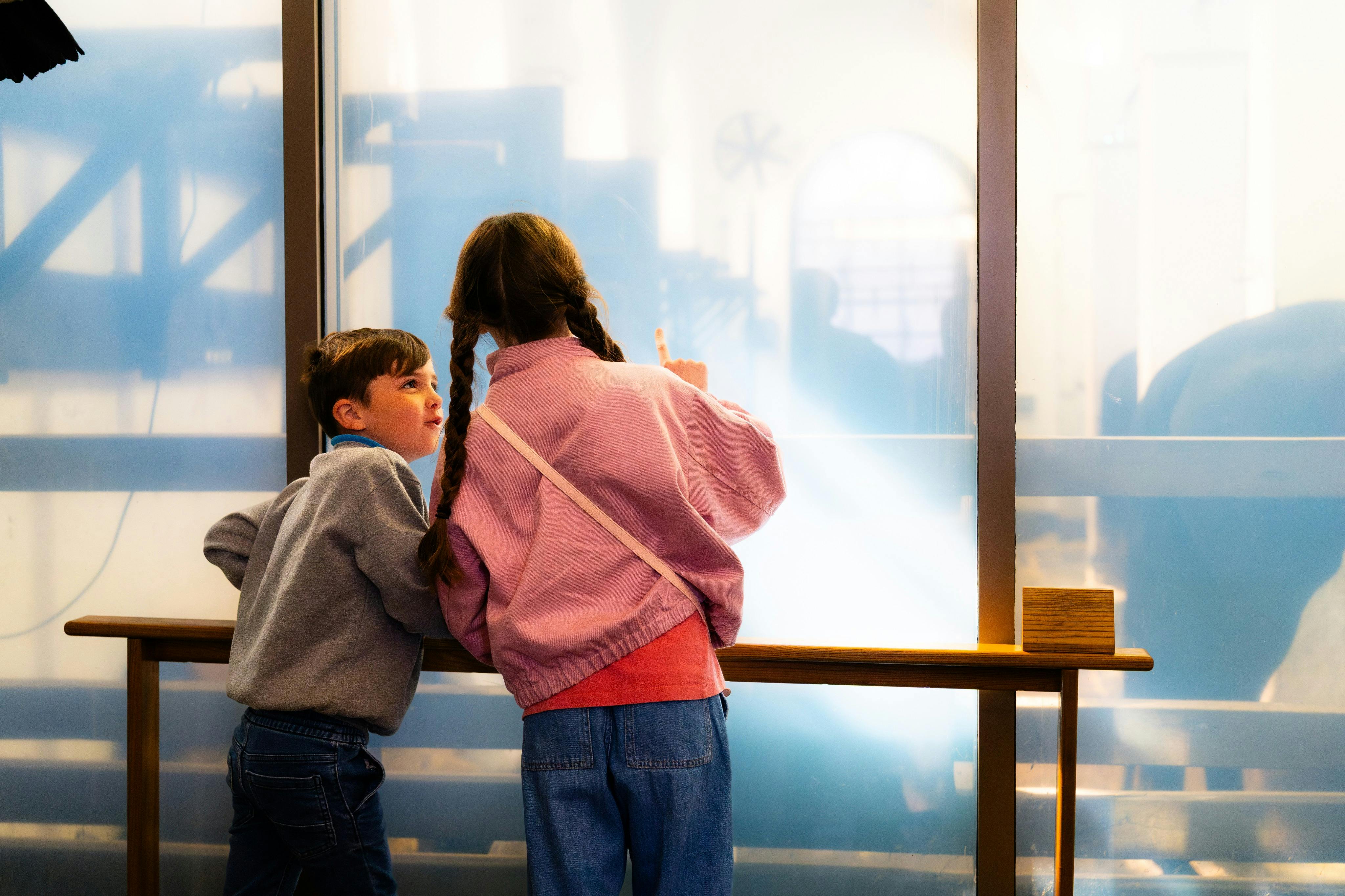 Dvě děti stojí u zábradlí, jedno ukazuje na průsvitné okno se strojem viditelným v pozadí.