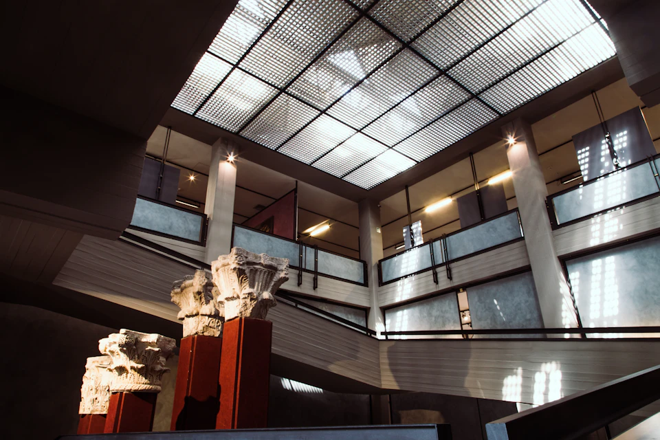 An interior with high ceilings, grid-patterned skylight, Greek columns, and multiple levels with metal railings.