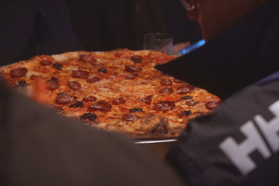 Close-up of a pepperoni pizza being served with blurred figures and a glass in the background.