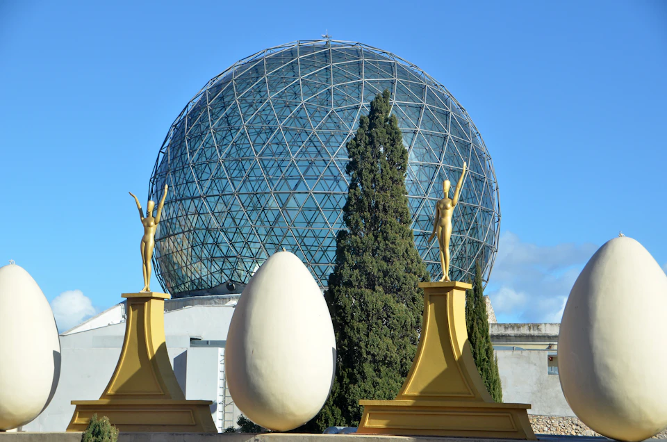 A geodesic dome with a grid pattern, flanked by statues and a large egg sculpture in front, with trees and a blue sky.