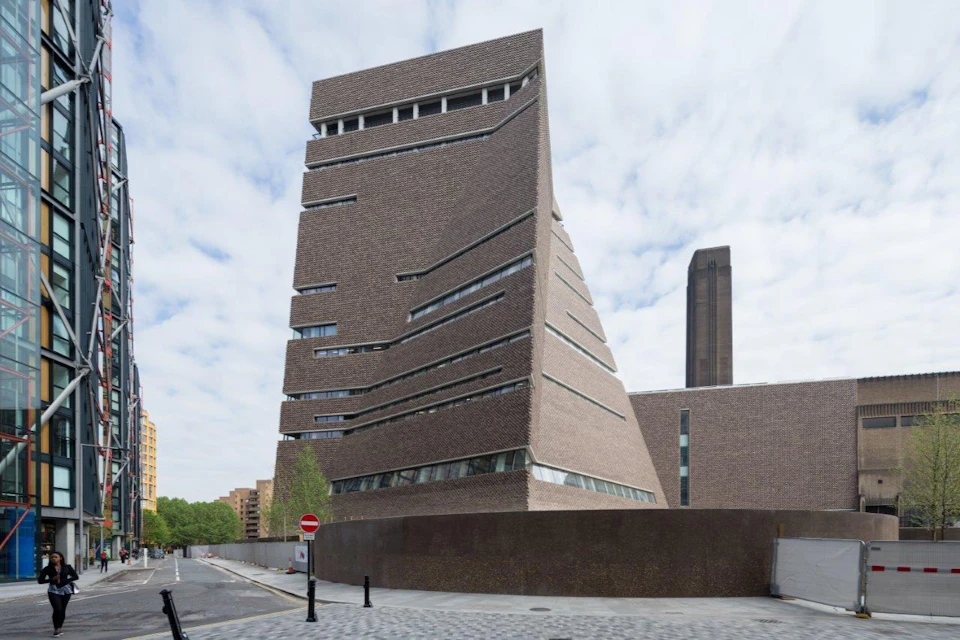 A modern, angular brick building with slanted walls and narrow windows, set against a partly cloudy sky.