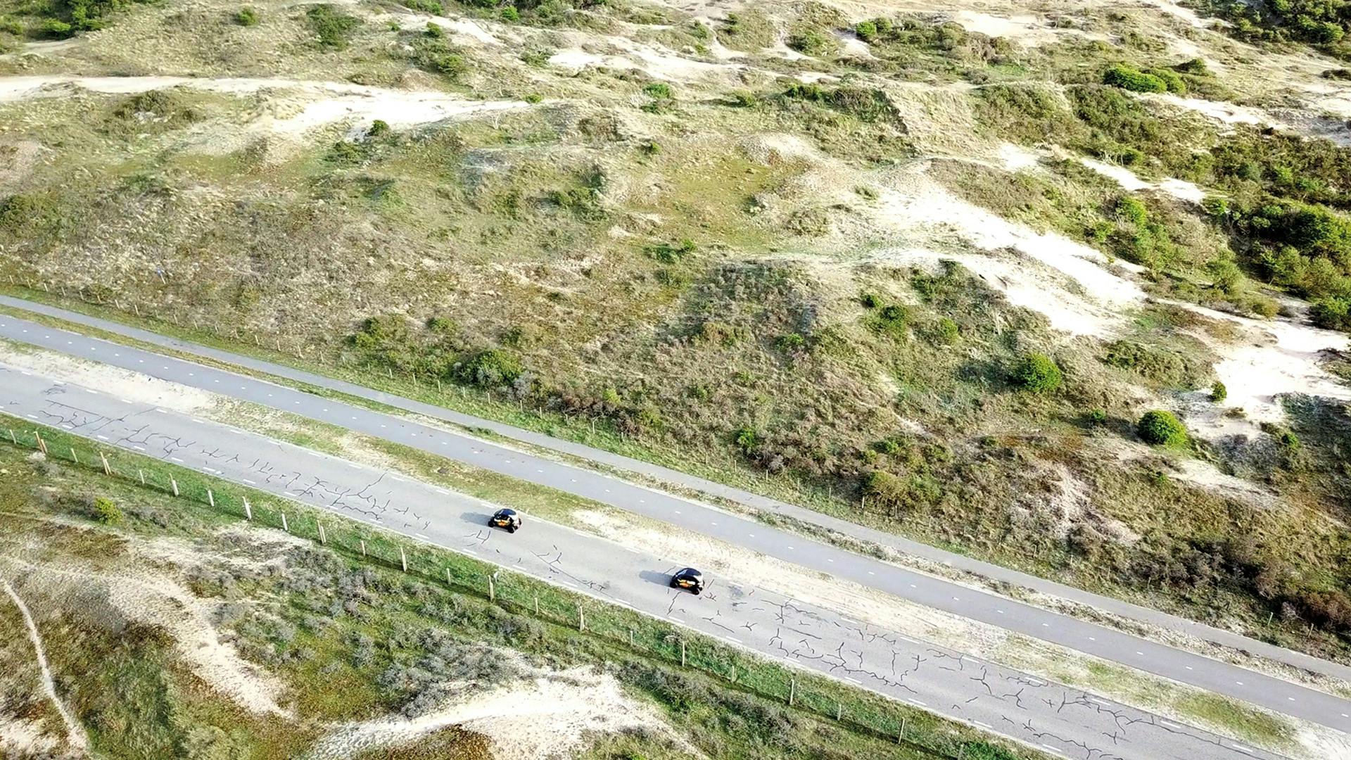 Luftfoto af to biler, der kører på en vej gennem et græsklædt, kuperet landskab med nogle spredte grønne områder.