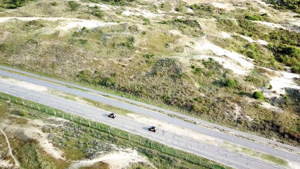 Aerial view of two cars driving on a road through a grassy, hilly landscape with some scattered patches of greenery.