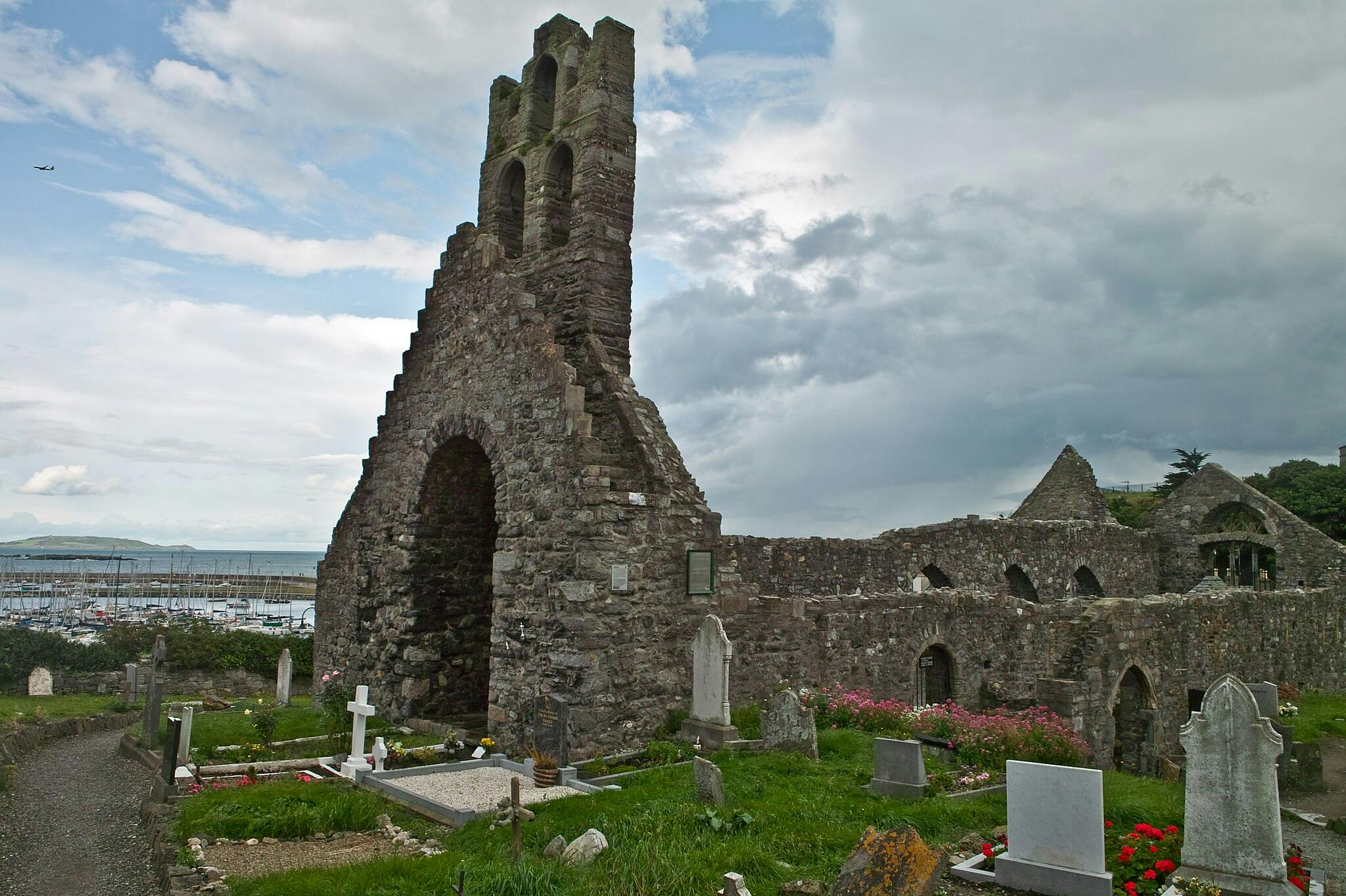 Old stone church ruins with arched windows and a tower, surrounded by a graveyard and flowers, near a harbor with many boats.