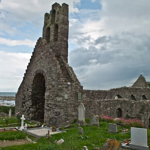 Gamla stenkyrkoruiner med välvda fönster och ett torn, omgivet av en kyrkogård och blommor, nära en hamn med många båtar.