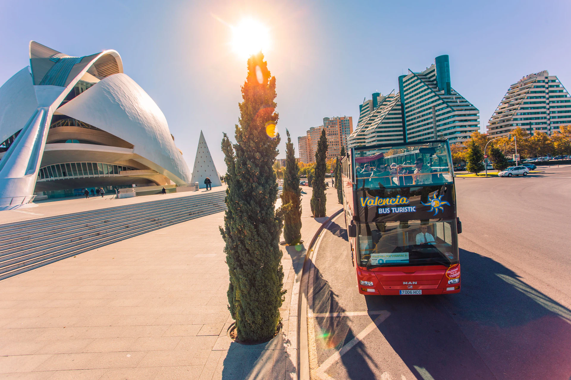 A red double-decker tourist bus drives on a sunny street lined with trees, near modern and futuristic buildings.