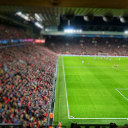 En fullsatt arena tittar på en fotbollsmatch på kvällen, med spelare i röda och vita uniformer på en starkt upplyst plan.