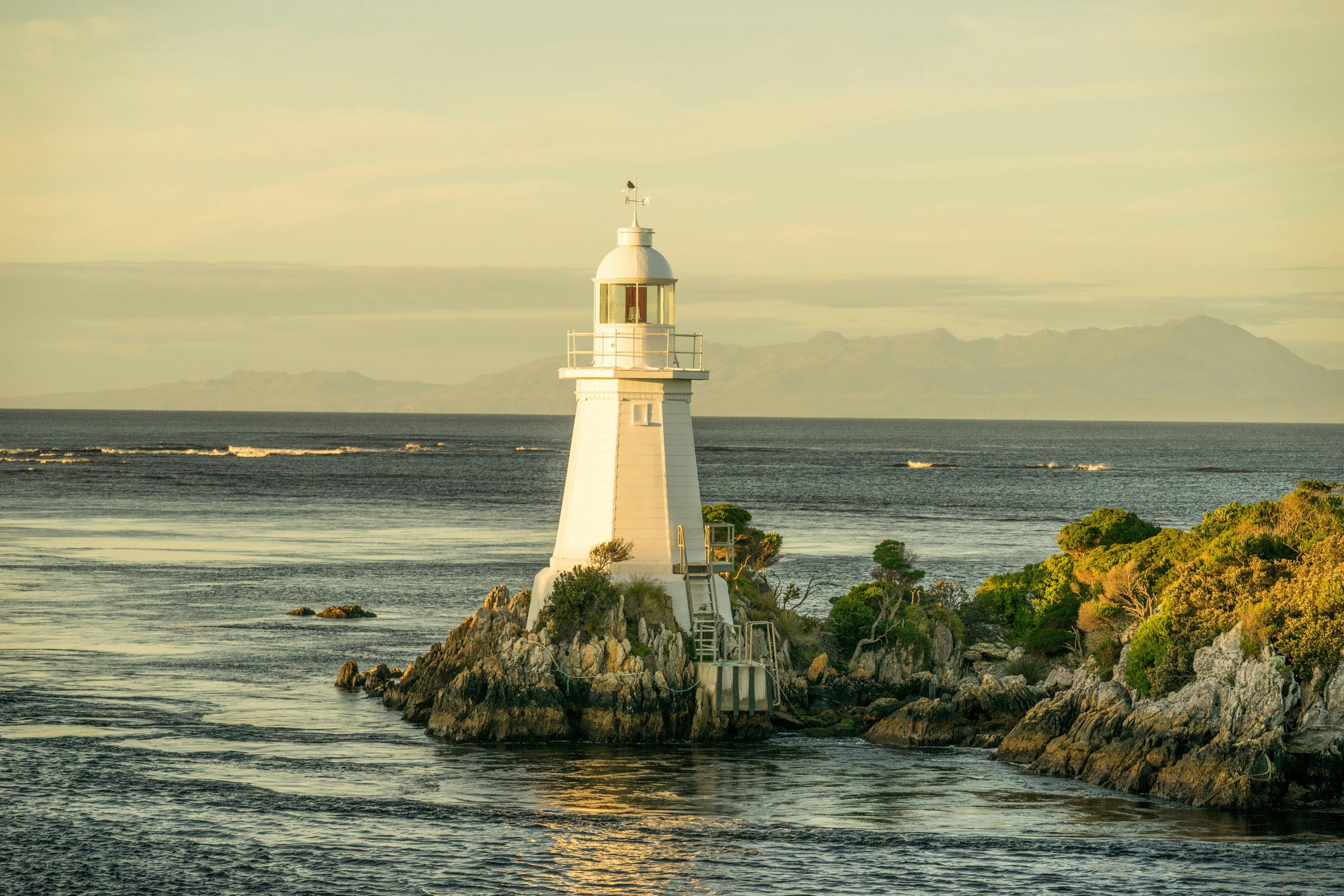 A white lighthouse on a small rocky island surrounded by calm water and greenery under a soft, golden sky.