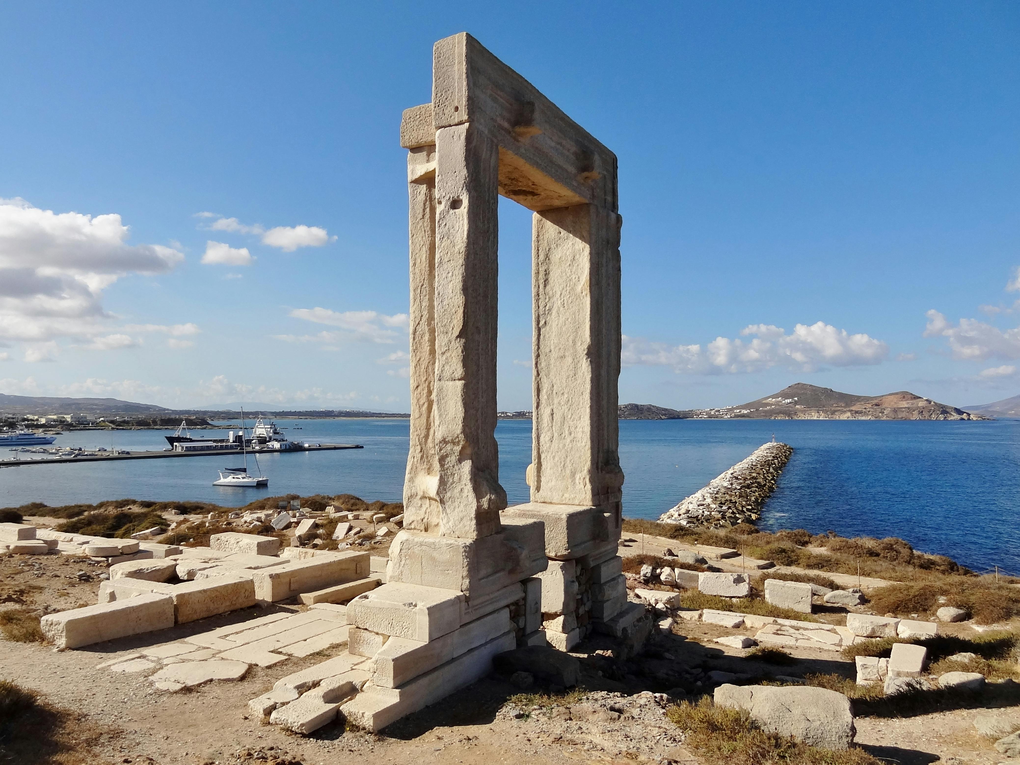 Ancient stone gateway ruins with rectangular frame, set against a coastal backdrop with calm blue water and distant mountains.