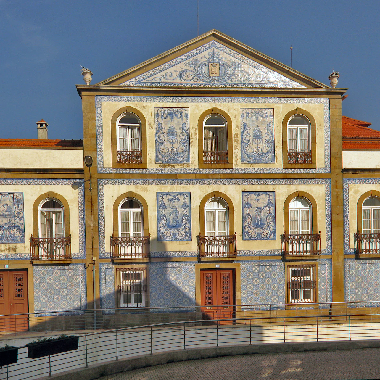 Ornate building with yellow walls, blue tile murals, arched windows with balconies, and a red-tiled roof against a clear sky.