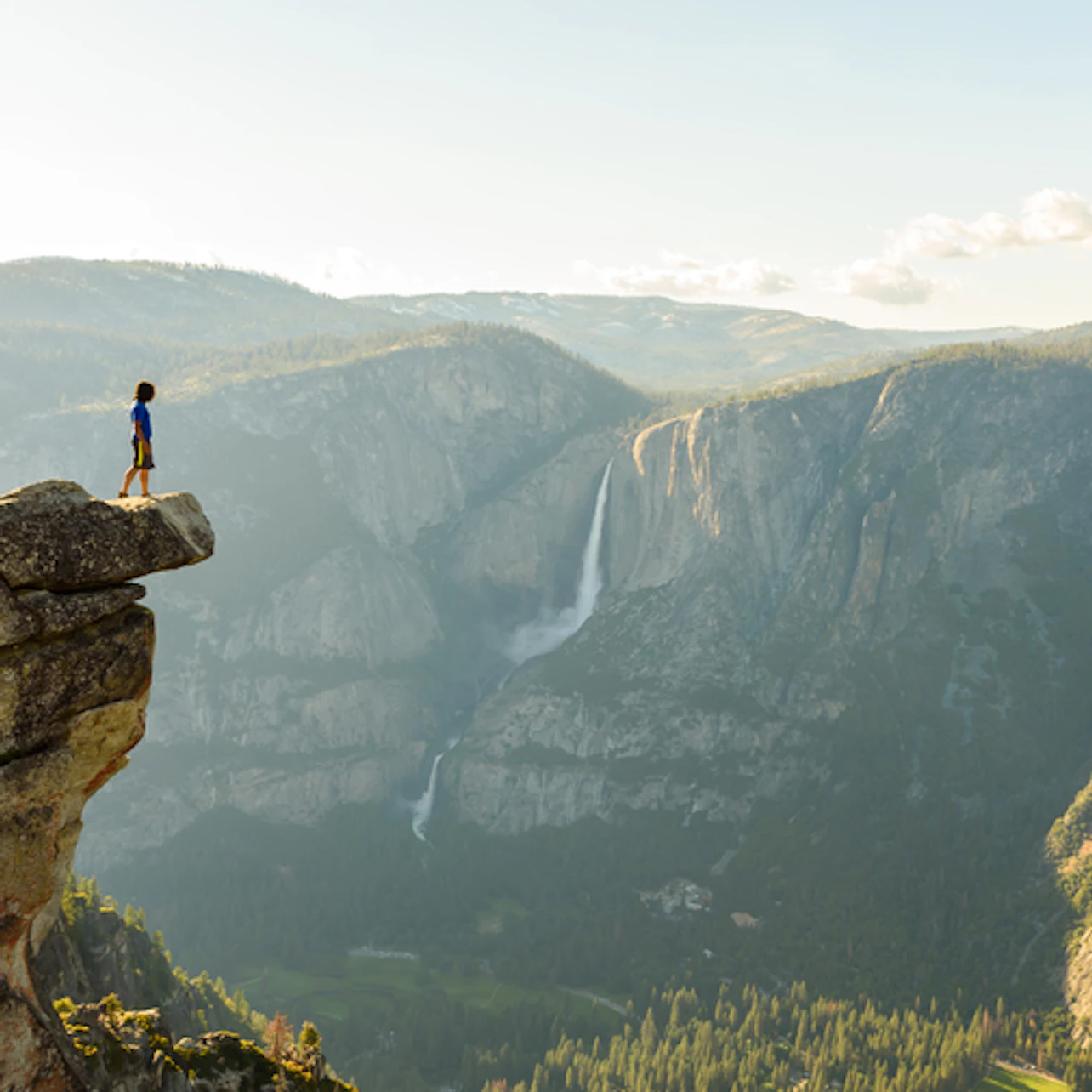 Person standing on a cliff edge overlooking a vast mountainous landscape with a distant waterfall.
