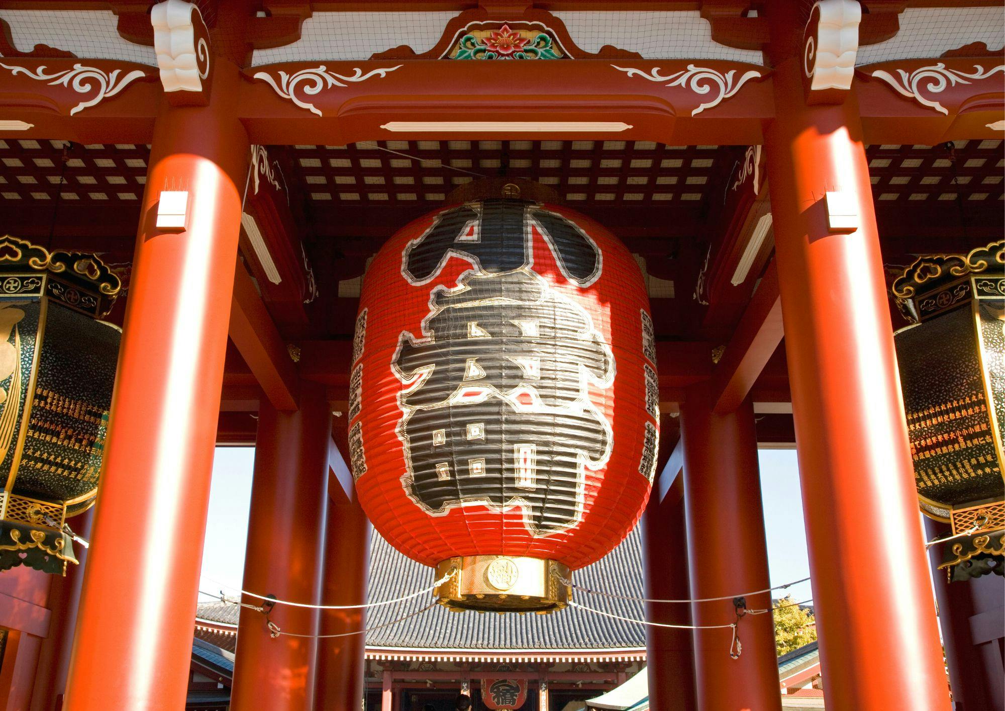 A large red lantern with black and white Japanese characters hangs in a traditional wooden temple gate.