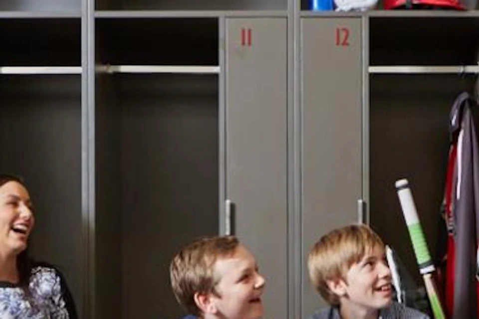 Four people sit on a bench in front of a row of lockers, conversing with a man in uniform holding a hand on one locker.
