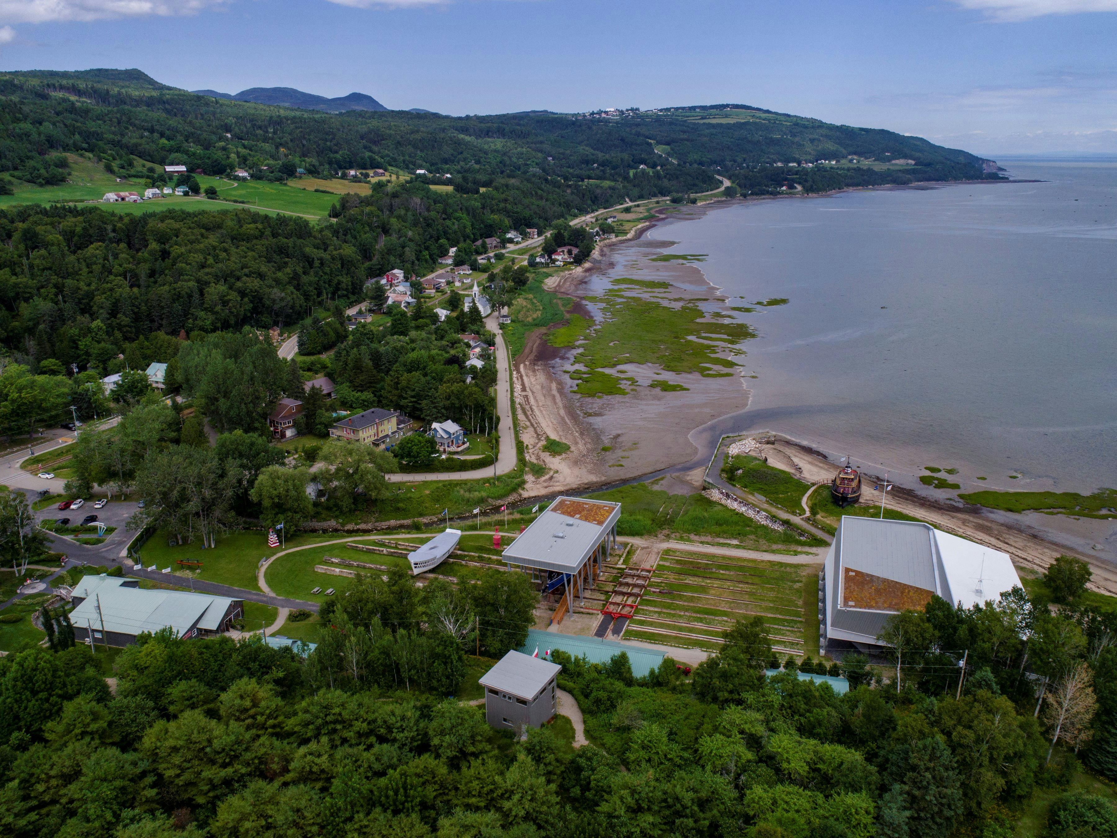 Luftfoto af en kystlandsby med bygninger, grønne bakker og en kystlinje med mudrede pletter ved vandet.
