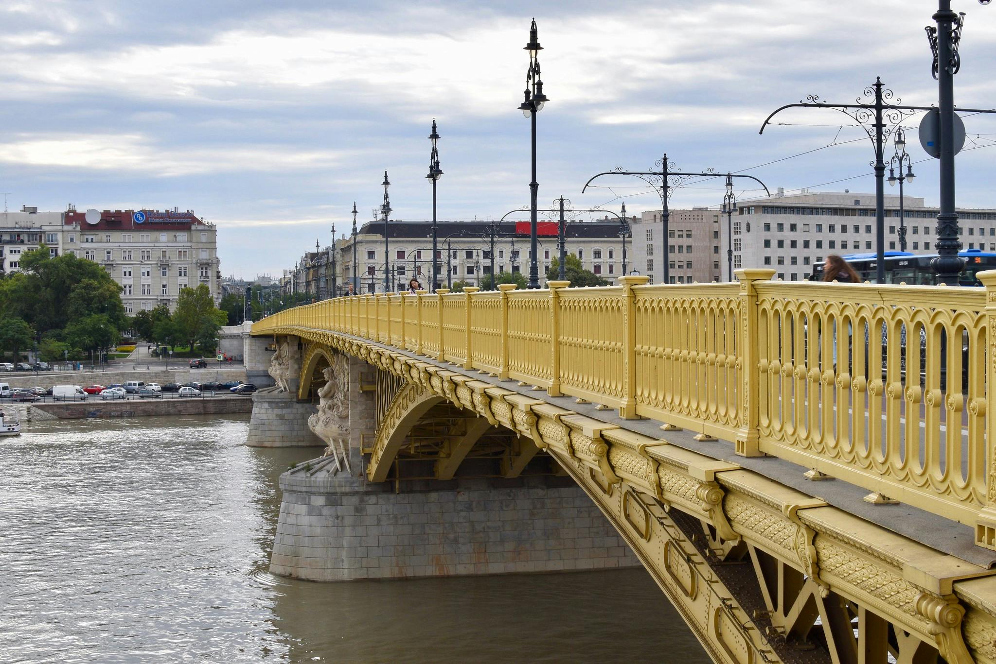 Gul bro med utsmyckade räcken sträcker sig över en flod. I bakgrunden finns byggnader, gatulampor och molnig himmel.