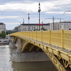 Gul bro med utsmyckade räcken sträcker sig över en flod. I bakgrunden finns byggnader, gatulampor och molnig himmel.