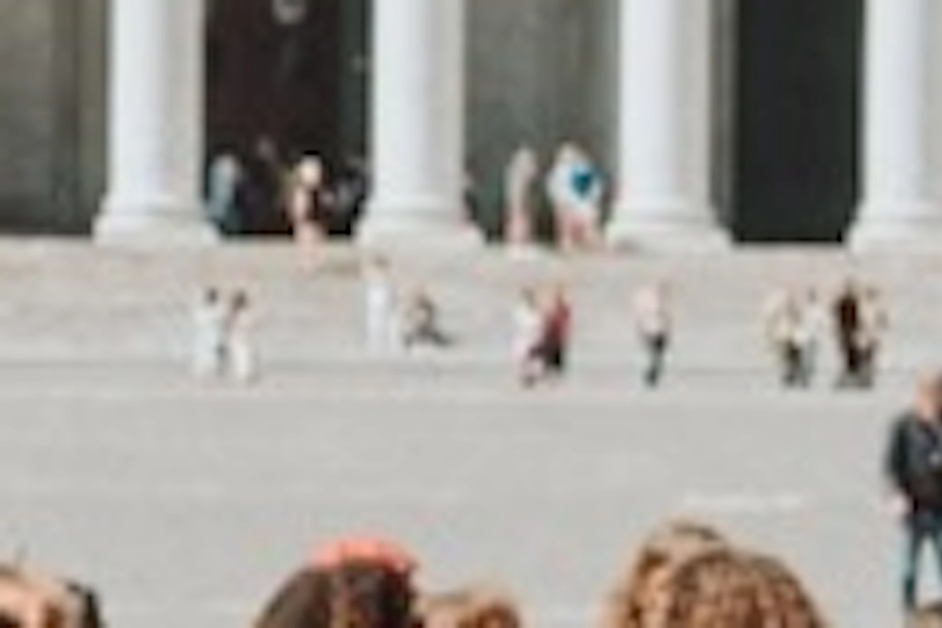 A group of people gathered in a square with a domed building and colonnades in the background under a blue sky.