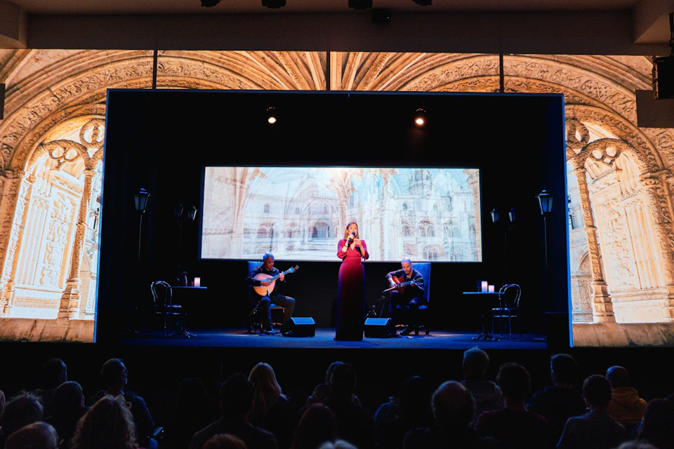 A woman in a red dress sings on stage with a guitarist on each side, set against a backdrop of a historic building image.