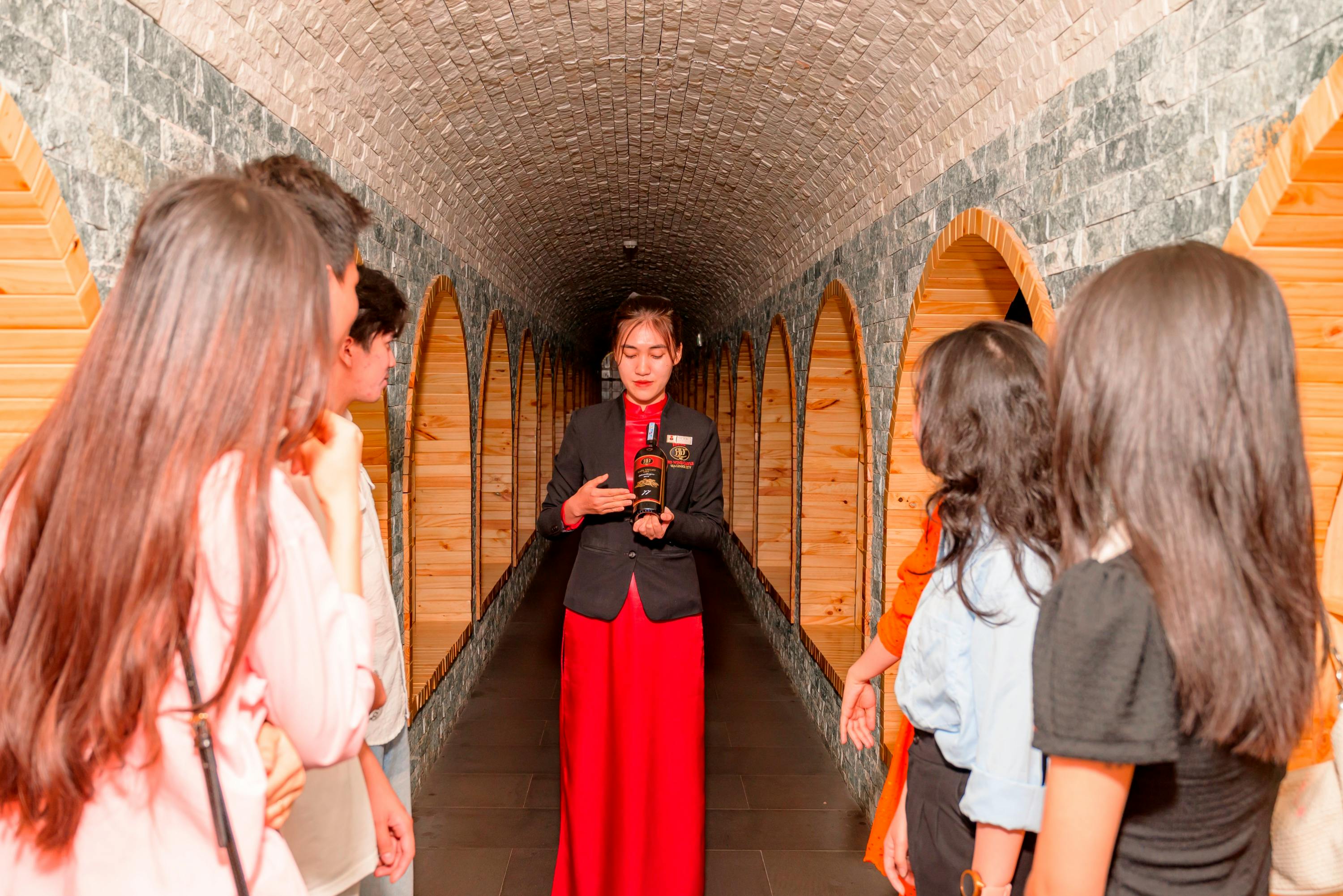 A guide in a red dress and black jacket explains to a group of people in a stone-walled tunnel lined with wooden arches.