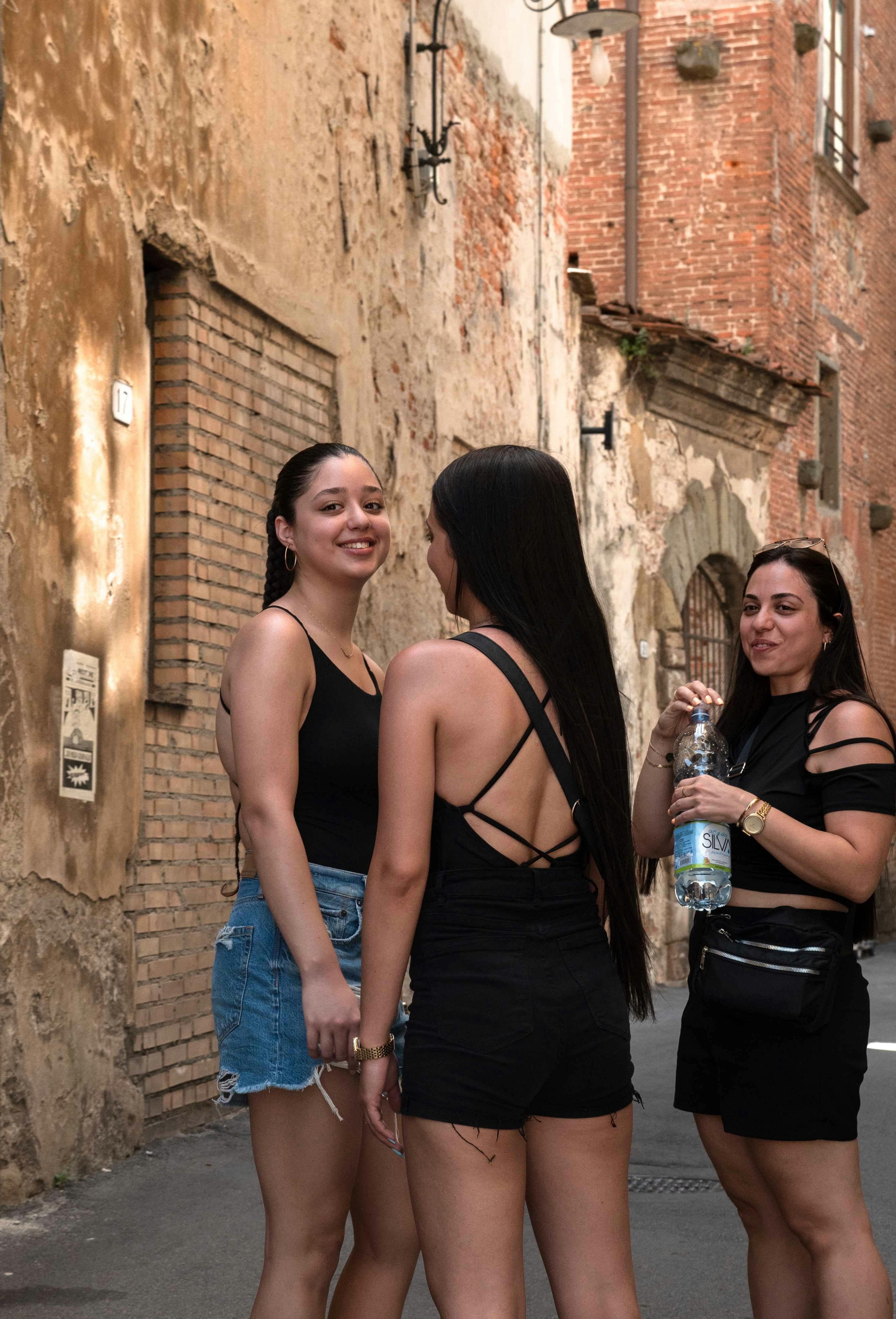 Three women stand chatting and smiling in a narrow street with weathered brick walls; one holds a bottle of water.