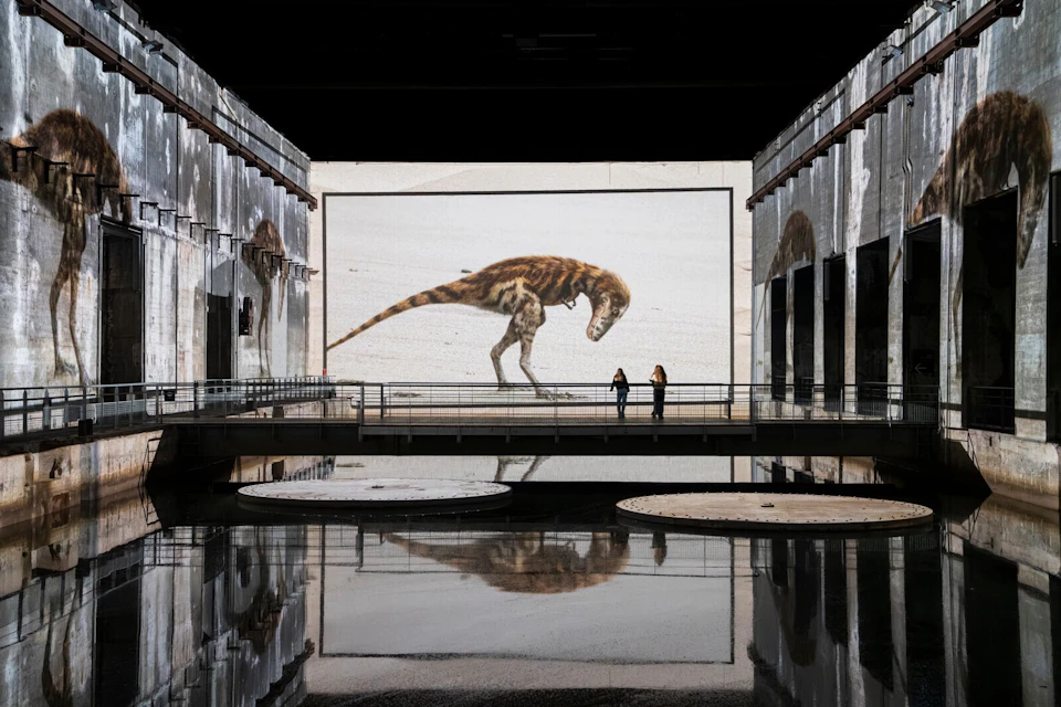 A large dinosaur image displayed on a screen in an industrial-style space with reflective water and two people observing.