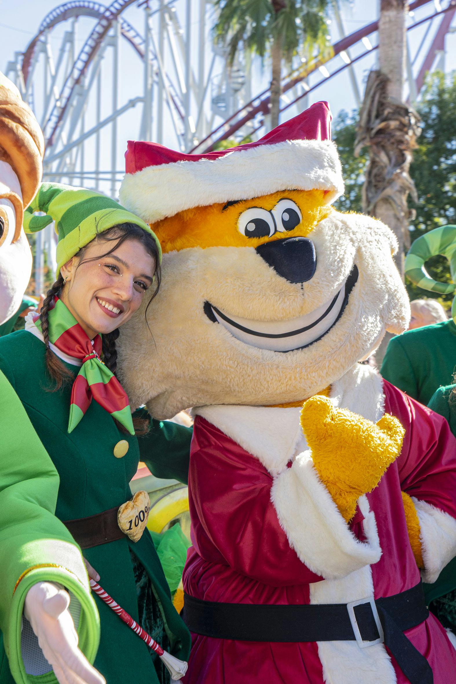 엘프 의상을 입은 사람이 산타 복장을 한 대형 마스코트 옆에서 테마파크를 배경으로 미소를 짓고 있습니다.